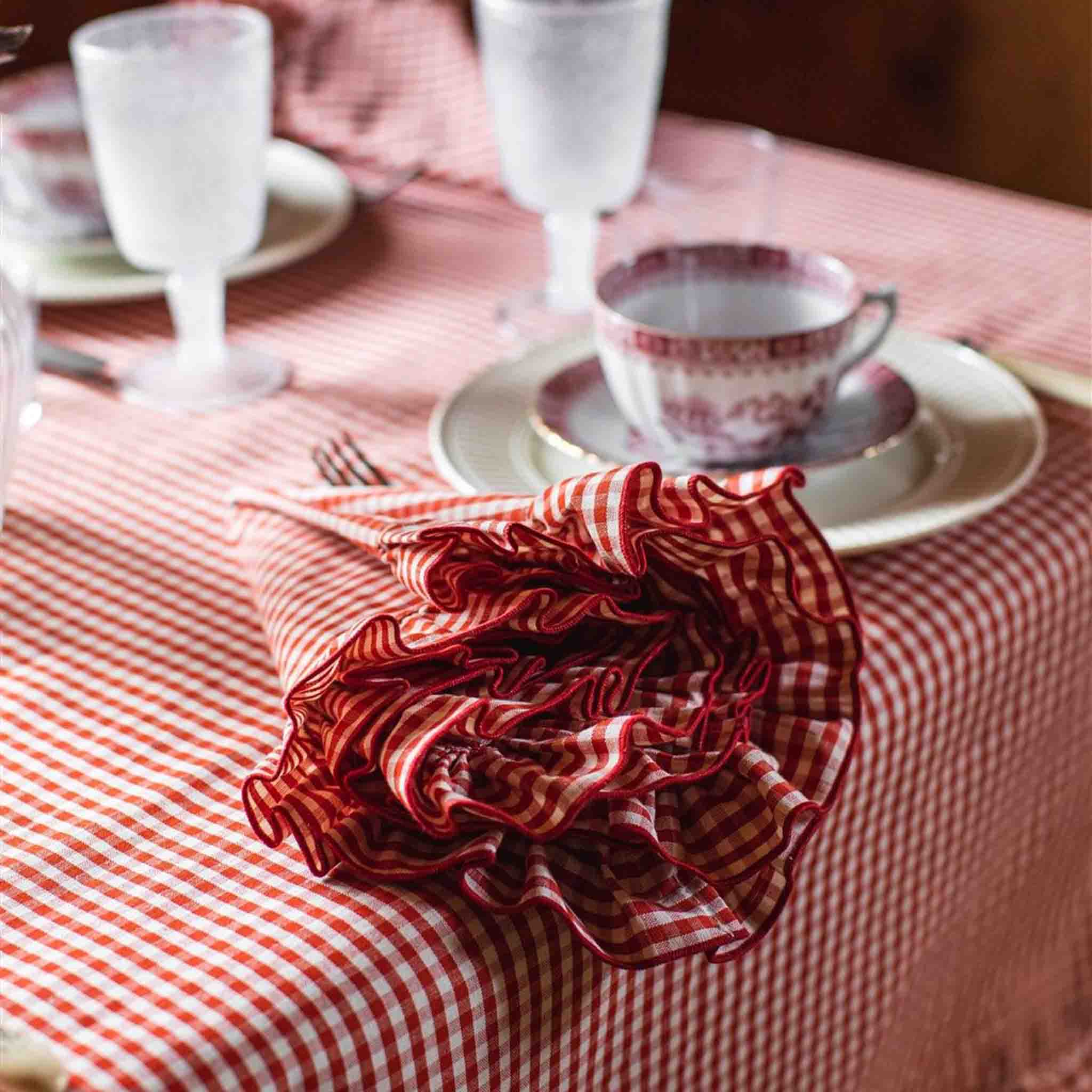 A red and white checkered napkin with ruffles placed on a table with a matching checkered tablecloth, with a cup and saucer in the background.