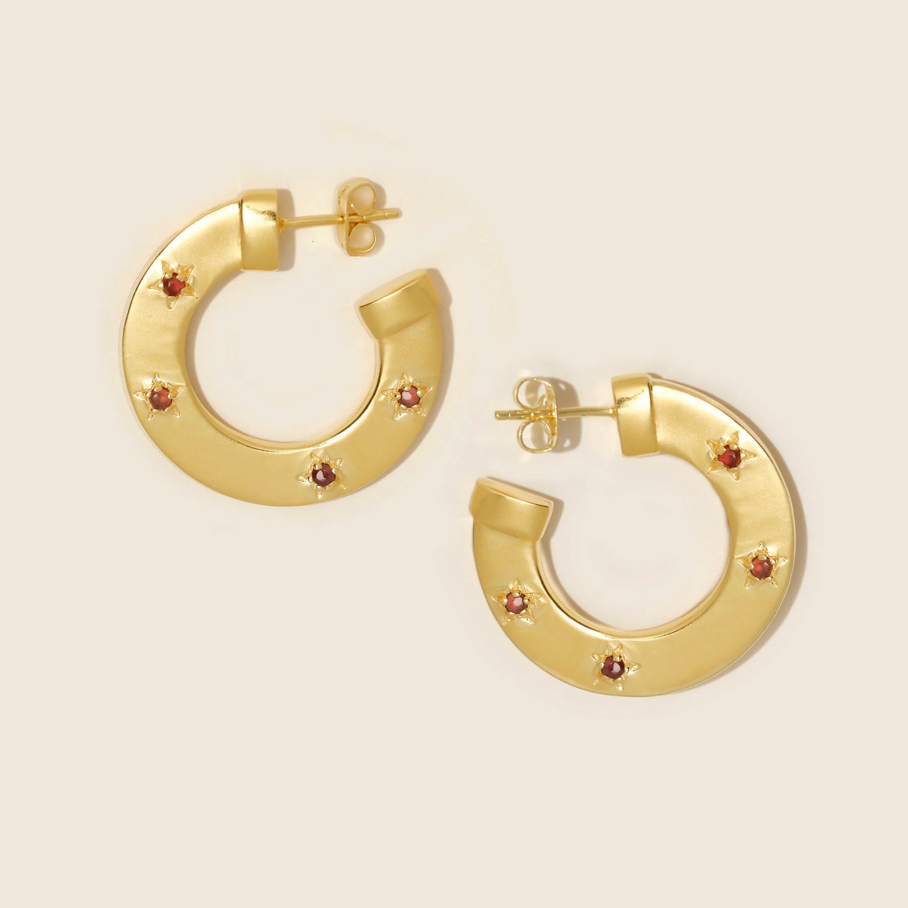 Gold hoop earrings with small red stones on a beige background
