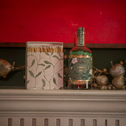 Botella Bottle Matches de Aura Collective junto a una caja de fósforos a juego con motivo de muérdago, sobre una repisa navideña con fondo rojo y decoración dorada.