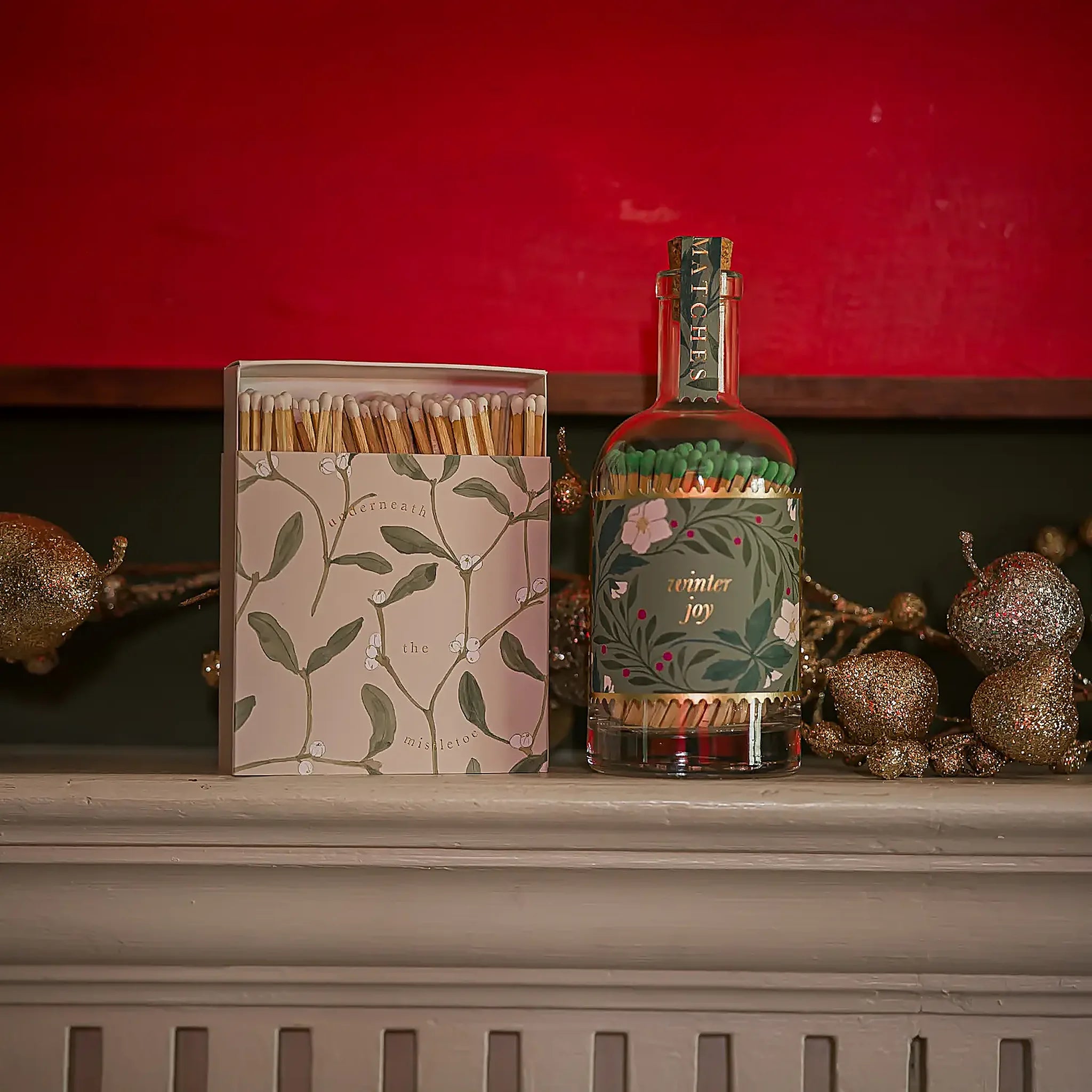 Botella Bottle Matches de Aura Collective junto a una caja de fósforos a juego con motivo de muérdago, sobre una repisa navideña con fondo rojo y decoración dorada.