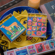 Caja de fósforos Colorful Gentleman de Aura Collective junto a otra caja de fósforos con un galgo, sobre un plato decorativo amarillo con forma de sol.