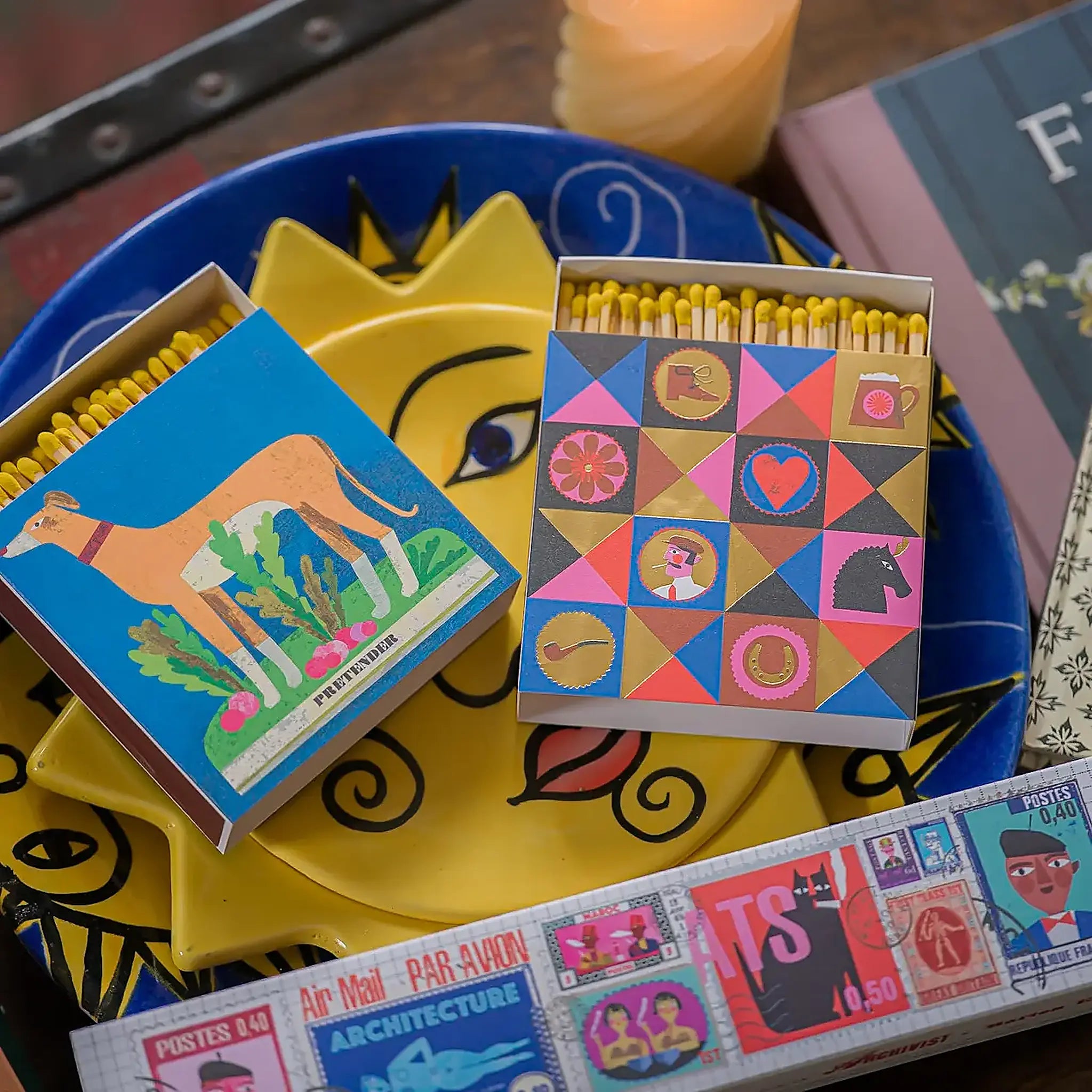 Caja de fósforos Colorful Gentleman de Aura Collective junto a otra caja de fósforos con un galgo, sobre un plato decorativo amarillo con forma de sol.