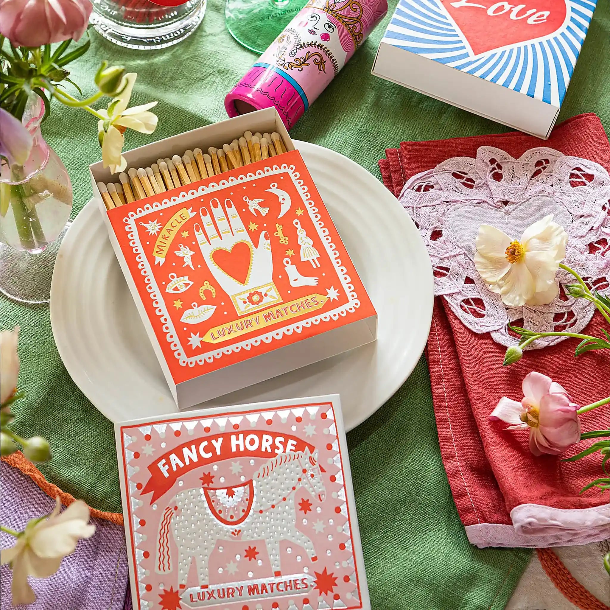 Caja de fósforos Fancy Horse de Aura Collective (diseño plateado de caballo sobre fondo rosa) en una mesa con flores, servilletas de encaje y una caja con un corazón que dice Love.