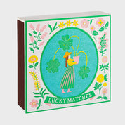 Foto de producto de la caja de fósforos Lucky Matches de Aura Collective: ilustración de una mujer con sombrero sosteniendo una maceta con tréboles de cuatro hojas, dentro de un óvalo celeste con marco floral verde y rosa.