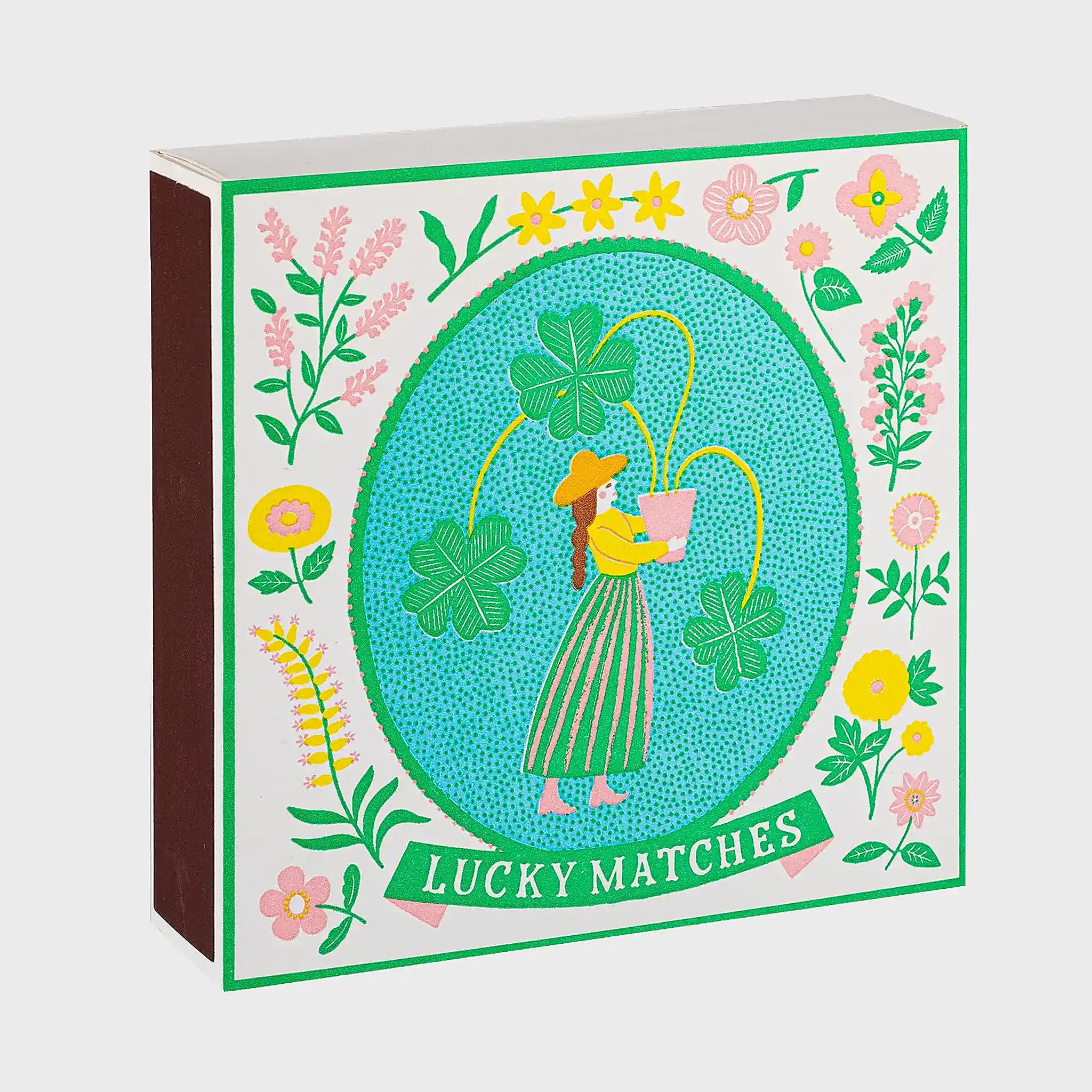 Foto de producto de la caja de fósforos Lucky Matches de Aura Collective: ilustración de una mujer con sombrero sosteniendo una maceta con tréboles de cuatro hojas, dentro de un óvalo celeste con marco floral verde y rosa.