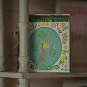 Caja de fósforos Lucky Matches de Aura Collective, con fósforos de punta verde asomando, sobre una repisa de madera blanca en un ambiente hogareño.