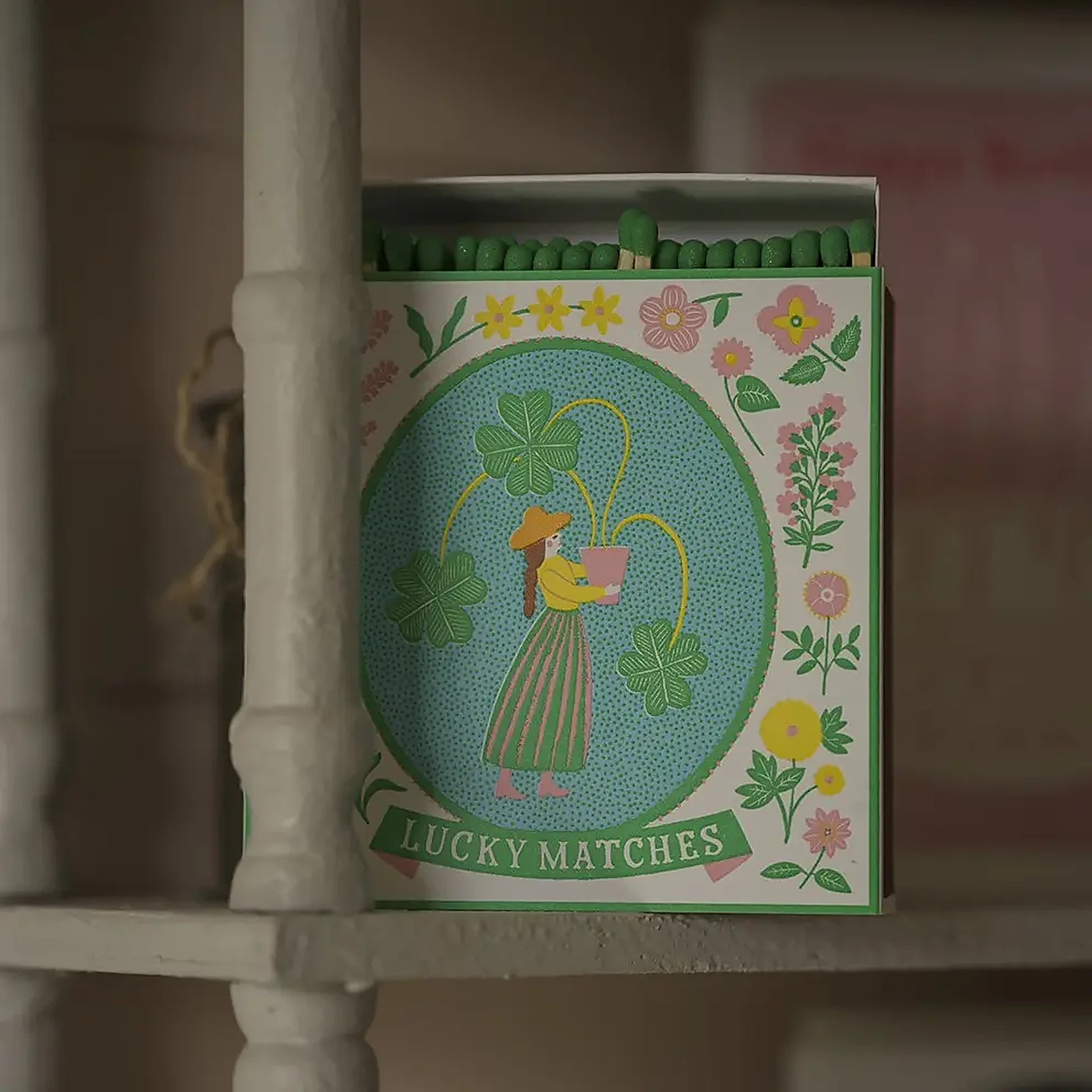Caja de fósforos Lucky Matches de Aura Collective, con fósforos de punta verde asomando, sobre una repisa de madera blanca en un ambiente hogareño.
