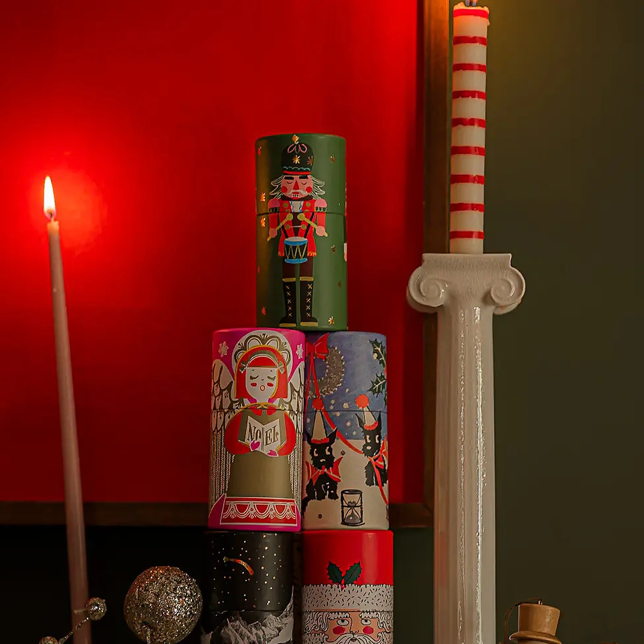 Stack of Nutcrackers Large Cylinder Matches tins in assorted holiday designs, styled with a lit taper candle