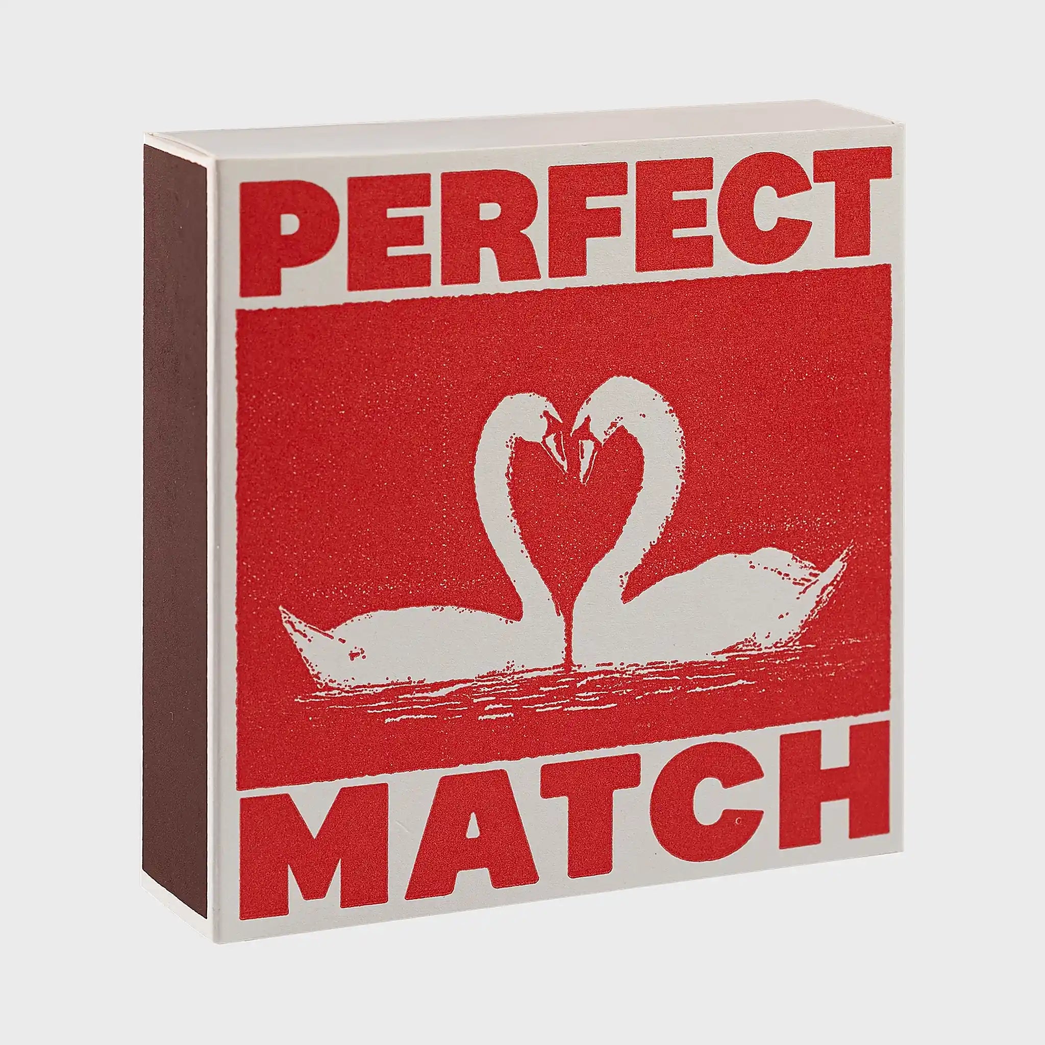 Foto de producto de la caja de fósforos Perfect Match de Aura Collective: fondo rojo con dos cisnes blancos formando un corazón con sus cuellos, y el texto Perfect Match.
