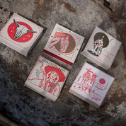 Caja de fósforos Saint No de Aura Collective junto a otras cajas de la misma colección con diseños de calavera de toro, caballo, ciclista y una pareja de vaqueros, sobre piedra.