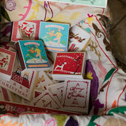 Caja de fósforos Season's Greetings de Aura Collective entre otras cajas de fósforos navideñas de diseño (Santa Claus, patinadores) sobre una tela bordada.