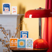 Caja de fósforos Sunflower de Aura Collective (diseño de girasol amarillo sobre fondo celeste) junto a otras tarjetas y cajas de diseño sobre una mesa, con una lámpara roja de fondo.