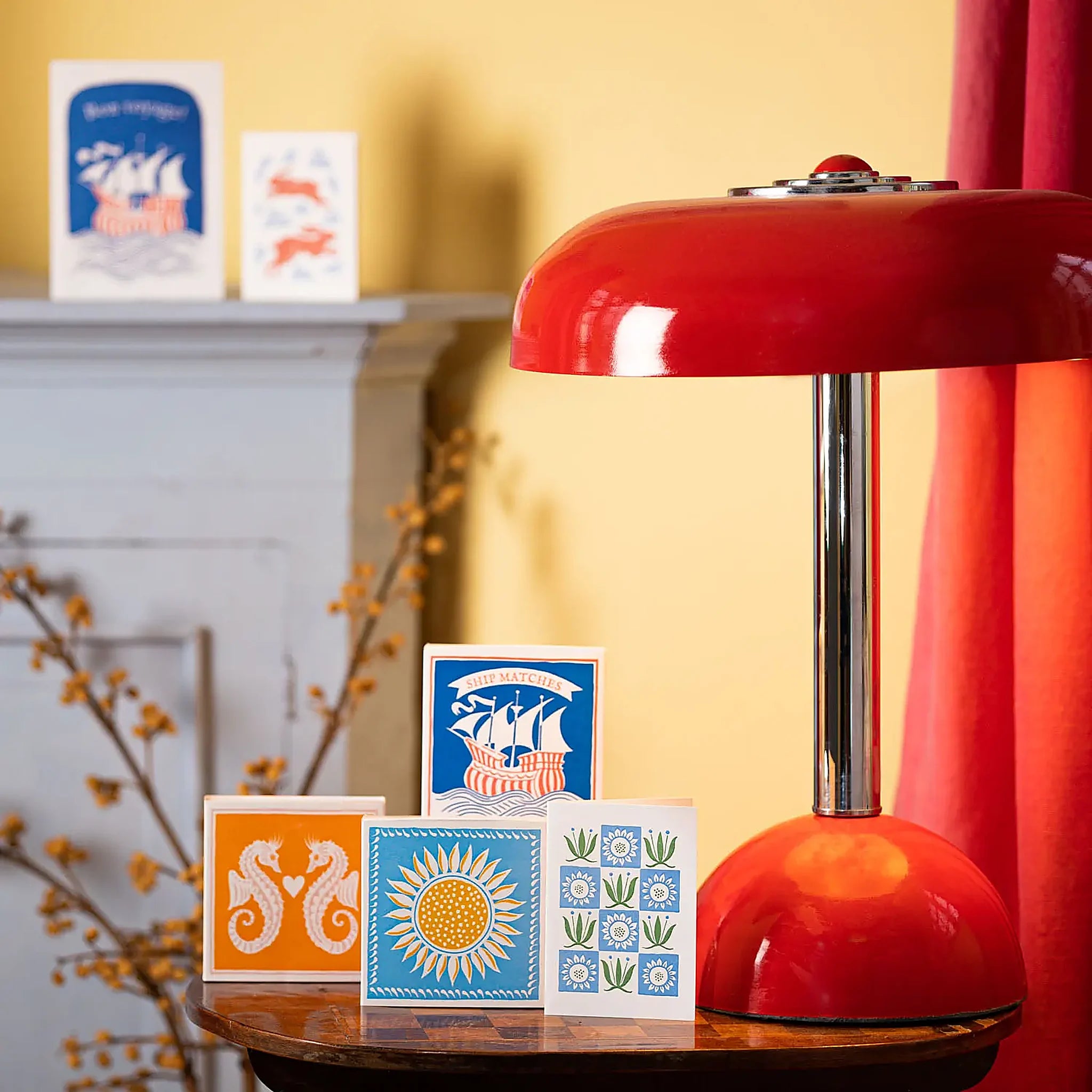 Caja de fósforos Sunflower de Aura Collective (diseño de girasol amarillo sobre fondo celeste) junto a otras tarjetas y cajas de diseño sobre una mesa, con una lámpara roja de fondo.