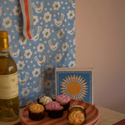 Caja de fósforos Sunflower de Aura Collective junto a una botella de vino y postres, con un mantel de fondo con estampado de pájaros azules.
