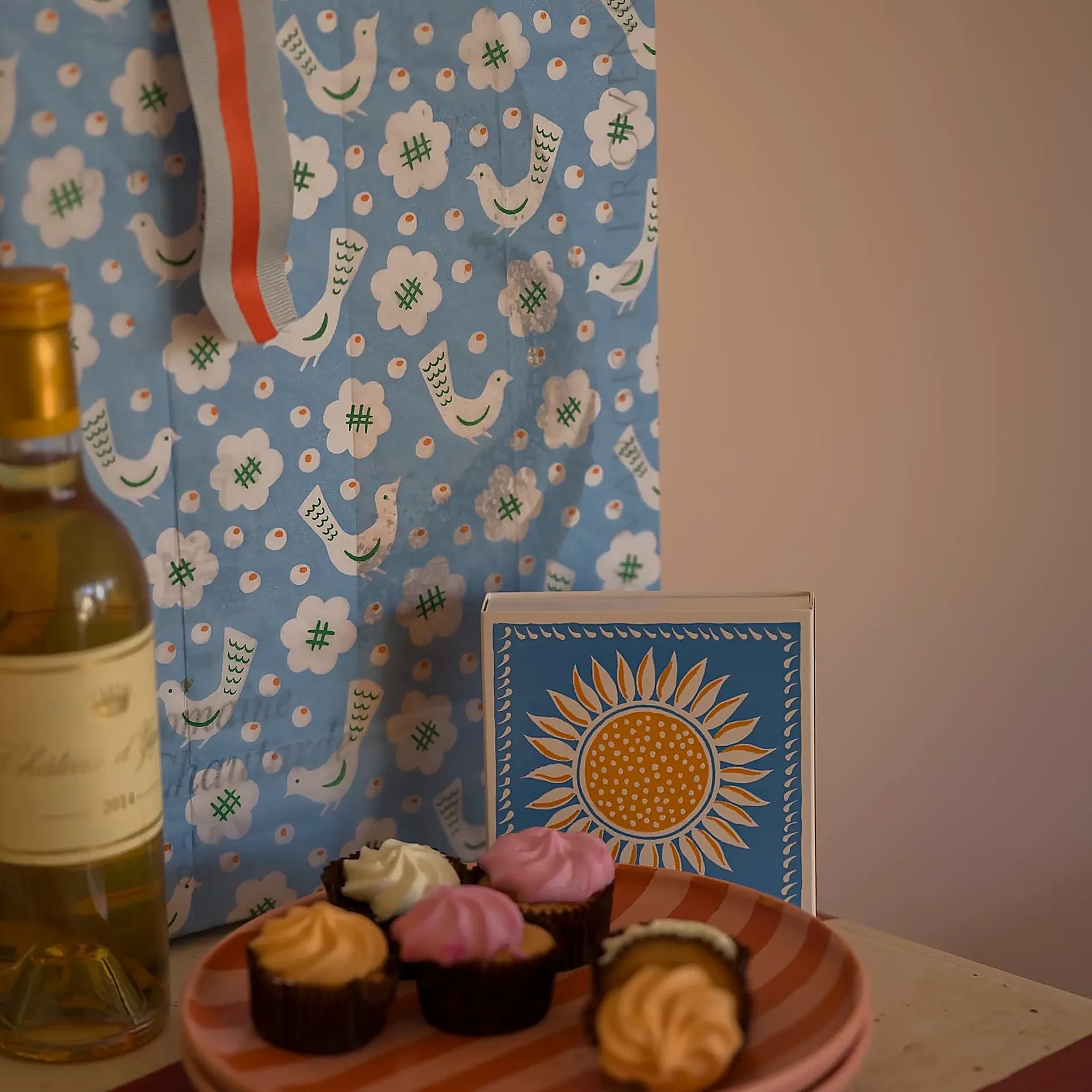 Caja de fósforos Sunflower de Aura Collective junto a una botella de vino y postres, con un mantel de fondo con estampado de pájaros azules.