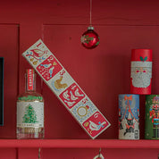 Caja de fósforos Twelve Days of Christmas de Aura Collective apoyada en una pared roja, junto a una botella de fósforos Merry Christmas y latas navideñas decorativas.