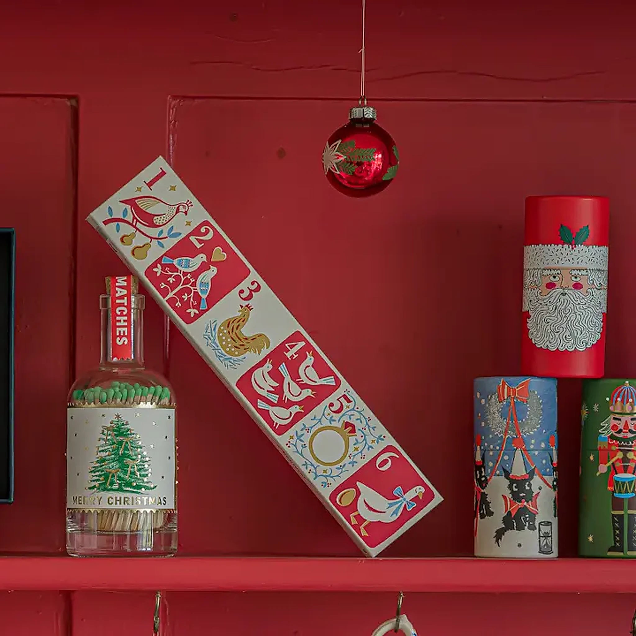 Caja de fósforos Twelve Days of Christmas de Aura Collective apoyada en una pared roja, junto a una botella de fósforos Merry Christmas y latas navideñas decorativas.